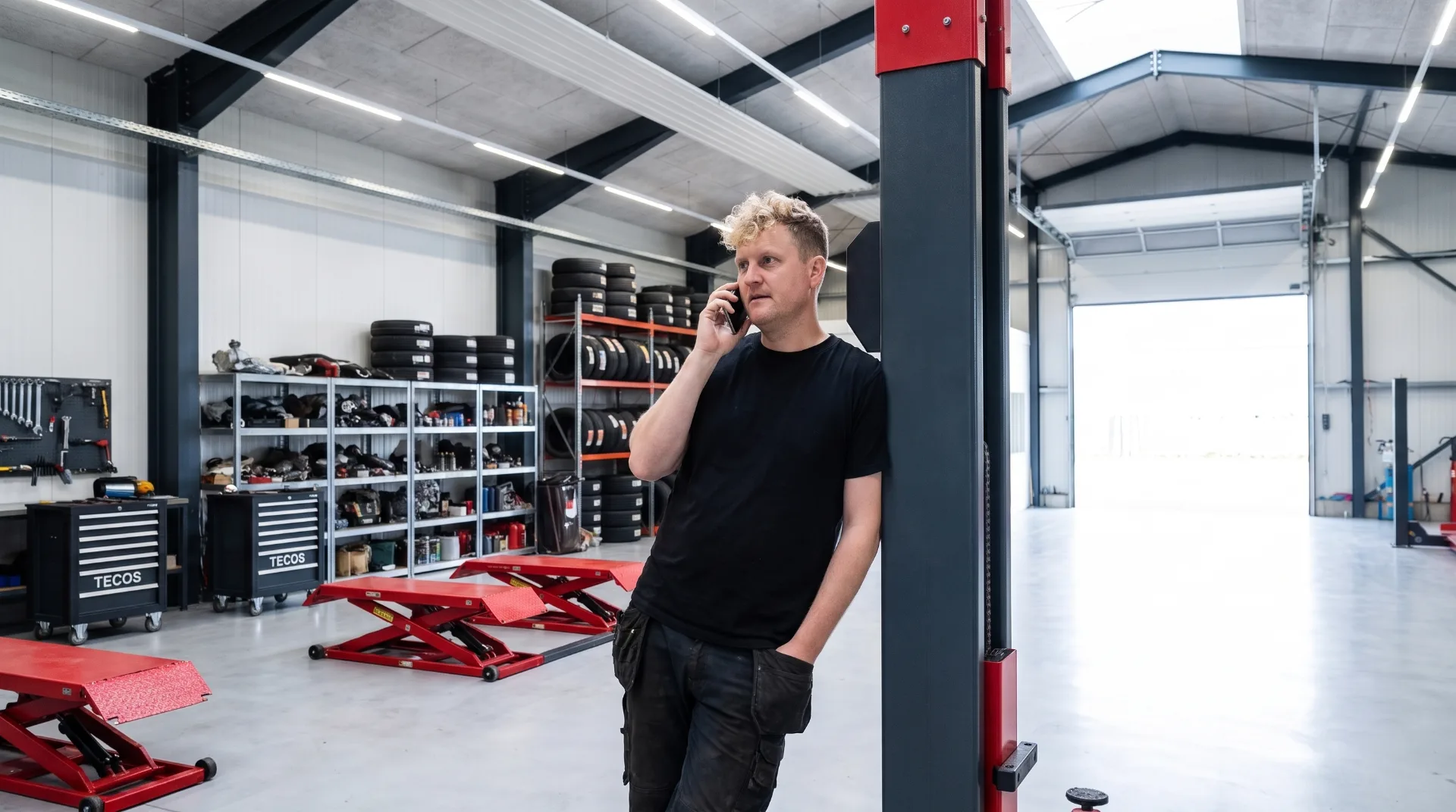 En mand i sort t-shirt læner sig op ad en lift-søjle og taler i telefon i det lyse værksted, med dæk på reoler og røde løfteborde i baggrunden